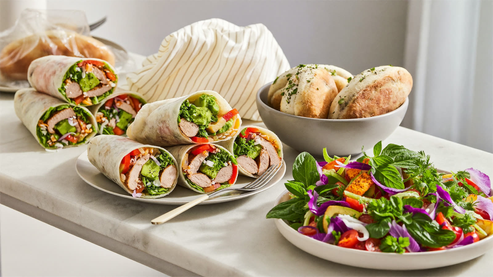 Fresh gourmet wraps cut open beside a rainbow garden salad bowl on a marble bench