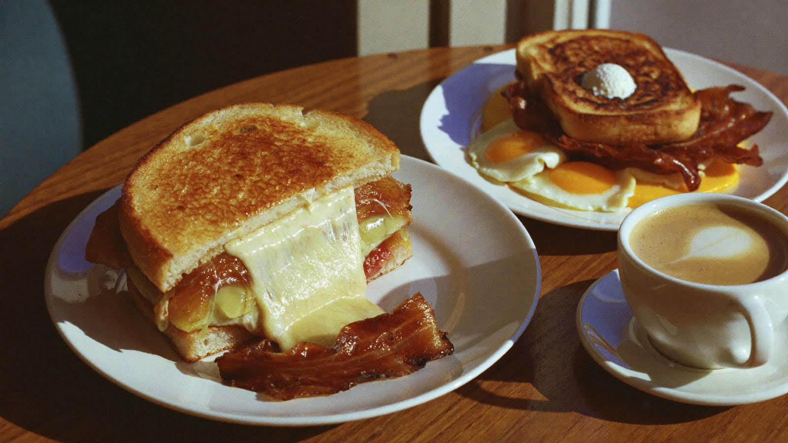 A golden toasted melt cut to show stretching cheese, with a breakfast plate of eggs and bacon and a flat white coffee